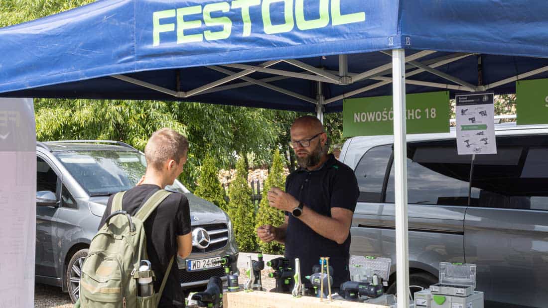 Doradca techniczny w okularach, ubrany w granatową koszulkę, stoi pod niebieskim namiotem z logo Festool. Rozmawia z młodą osobą, która ma na sobie czarny plecak.