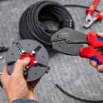 Szczypce KNIPEX MultiCrimp® PV trzymane w dłoni obok magazynka bębnowego z matrycami, w tle zwój kabla.