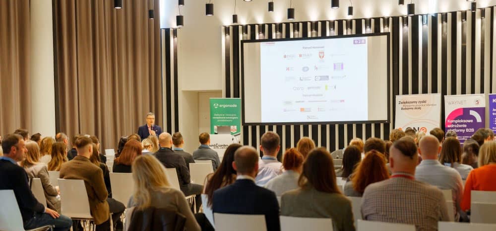Prelegent na scenie przemawia do pełnej sali uczestników Konferencji E-commerce B2B, na ekranie lista partnerów wydarzenia.