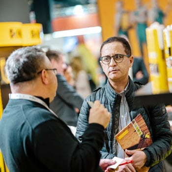 Uczestnik targów EISENWARENMESSE rozmawia z wystawcą przy stoisku z produktami budowlanymi/chemią w tubach.