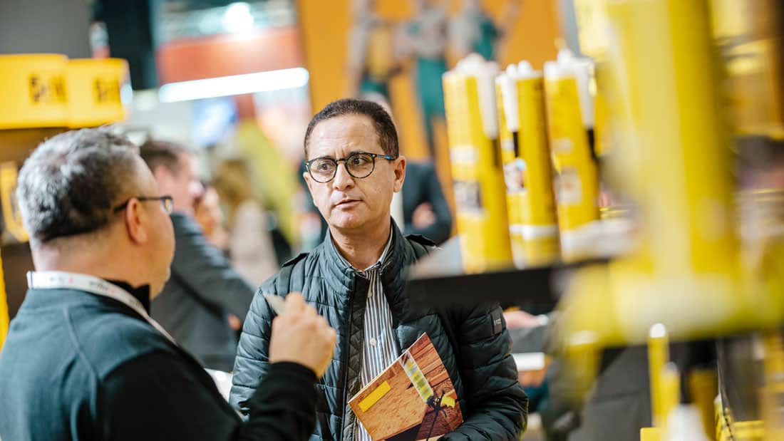 Uczestnik targów EISENWARENMESSE rozmawia z wystawcą przy stoisku z produktami budowlanymi/chemią w tubach.