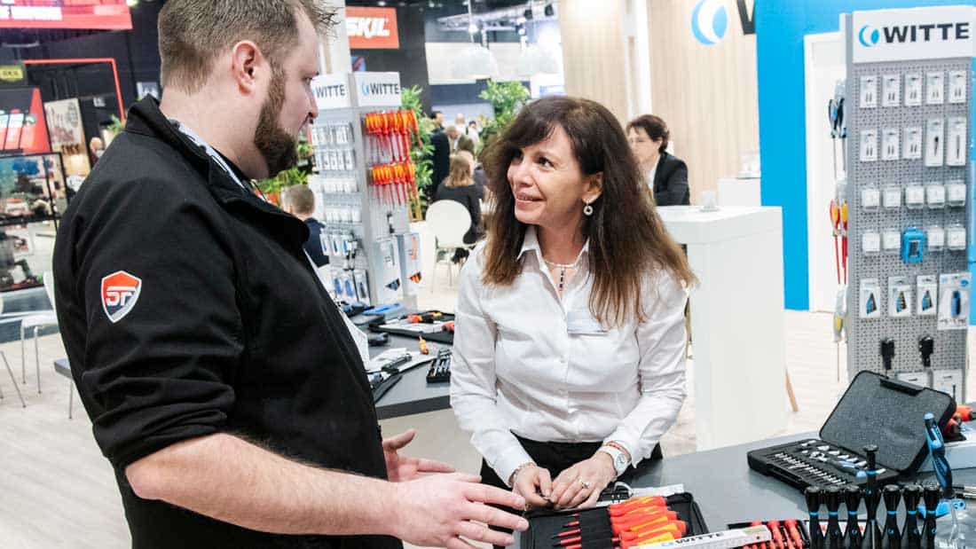 Wystawczyni rozmawia z klientem przy ekspozycji zestawów narzędzi i akcesoriów mocujących na targach EISENWARENMESSE.