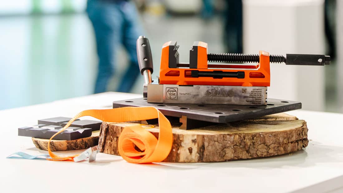 Detal pomarańczowego imadła maszynowego VERSAMAG (zacisk, element mocujący) i pomarańczowej taśmy na drewnianym podłożu, produkt z oferty targów International Hardware Fair.