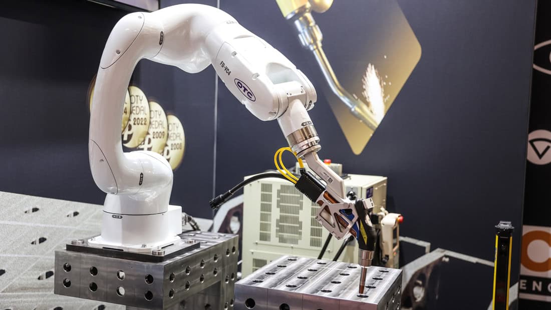 Biały robot przemysłowy OTC na stanowisku do spawania lub zgrzewania metalu. Detal maszyny z widocznymi nagrodami w tle.