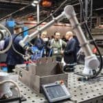 Robot przemysłowy w akcji na stanowisku spawalniczym, prezentujący możliwości automatyzacji obróbki metalu. Widoczna dyskusja biznesowa w tle.