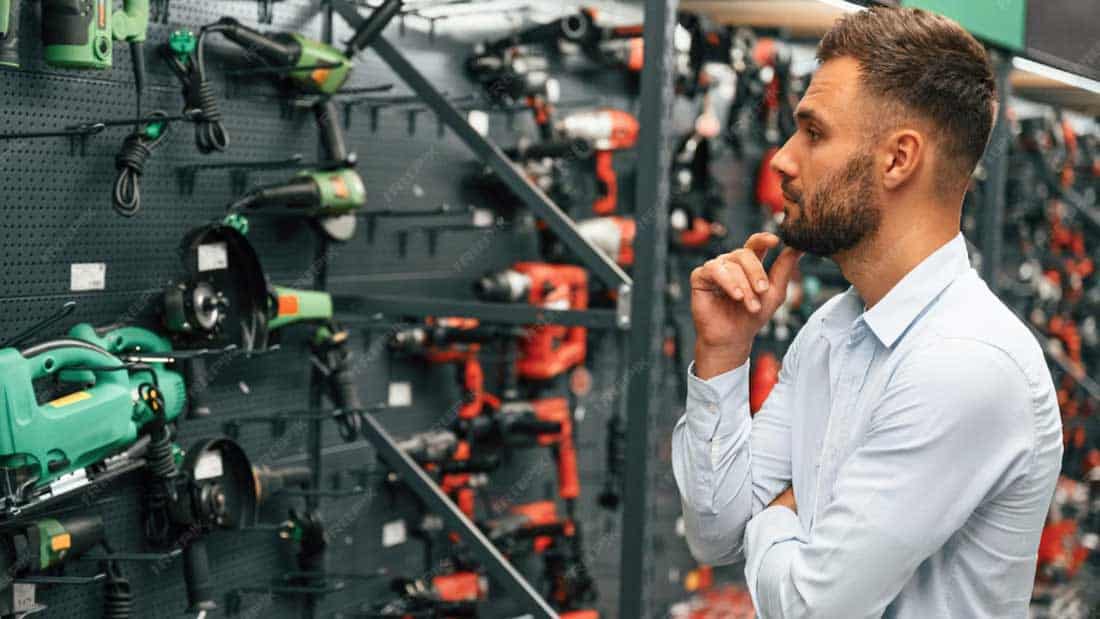 Mężczyzna w niebieskiej koszuli zastanawiający się nad wyborem profesjonalnych elektronarzędzi w dobrze zaopatrzonej hurtowni.