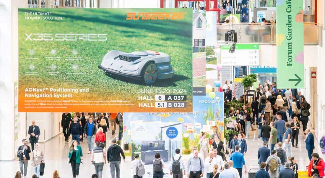 Panoramiczne ujęcie wnętrza targów spoga+gafa w Kolonii, ukazujące tłumy odwiedzających. Na pierwszym planie duży baner z reklamą robota koszącego SUNSEEKER X35 SERIES i informacjami o halach 6 i 5.1. Widoczny także zielony szyld Forum Garden Café. (Targi ogrodnicze, kosiarki automatyczne, tłum, Koelnmesse)