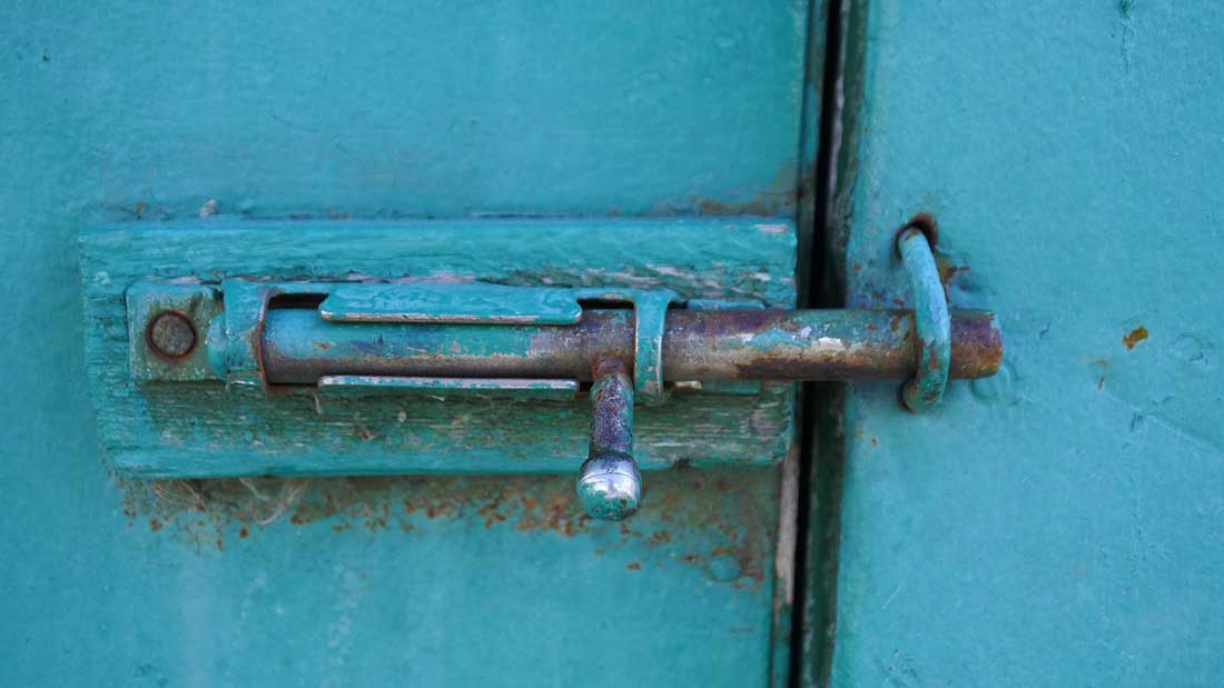 Metalowa zasuwa montowana na turkusowych drzwiach, z widocznymi oznakami zużycia i korozji