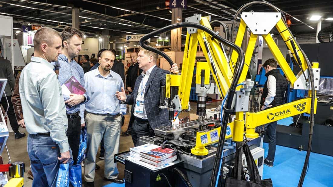 Pokaz działania żółtego robota przemysłowego CMA Machine Tools na stoisku targowym. Przedstawiciel wystawcy prezentuje możliwości automatyzacji i gwintowania grupie zainteresowanych inżynierów.