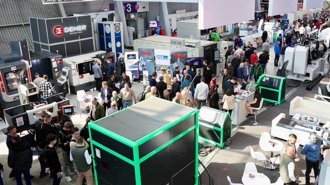 Zatłoczona hala targowa podczas targów STOM w Kielcach. W centrum kadru stoisko Dener Machine Tools oraz duże maszyny do obróbki metalu w kolorze zielonym i białym. Inżynierowie i specjaliści prowadzą rozmowy przy eksponatach.