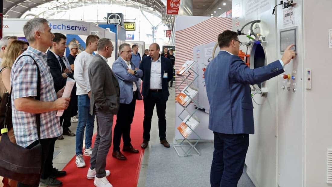 Prezentacja przemysłowego urządzenia do obróbki cieplnej lub testów materiałowych podczas targów w Targach Kielce; demonstracja panelu sterowania i rozmowy przedstawicieli branży metalurgicznej na stoisku wystawcy.