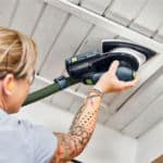 Akumulatorowa szlifierka delta Festool DTSC 200 podczas szlifowania drewnianego sufitu z podłączonym odkurzaczem i pierścieniem LED oświetlającym powierzchnię.